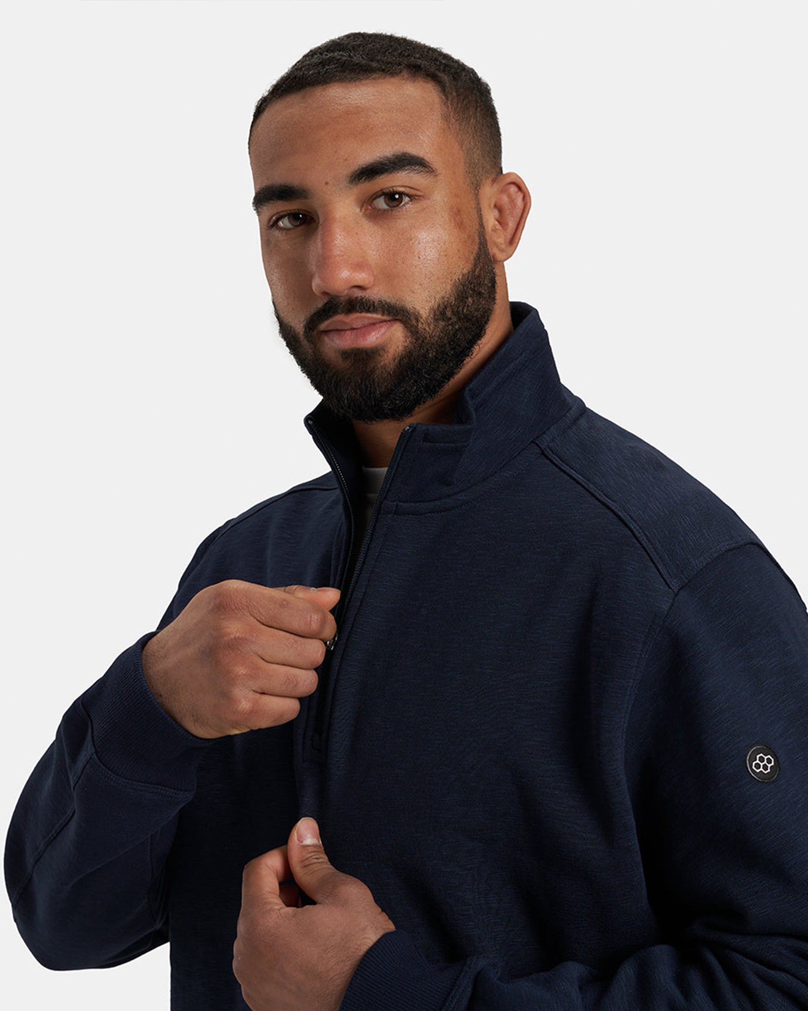 A close-up portrait of a man wearing a navy zip-up sweatshirt presenting a confident expression and adjusting the zip with his hands