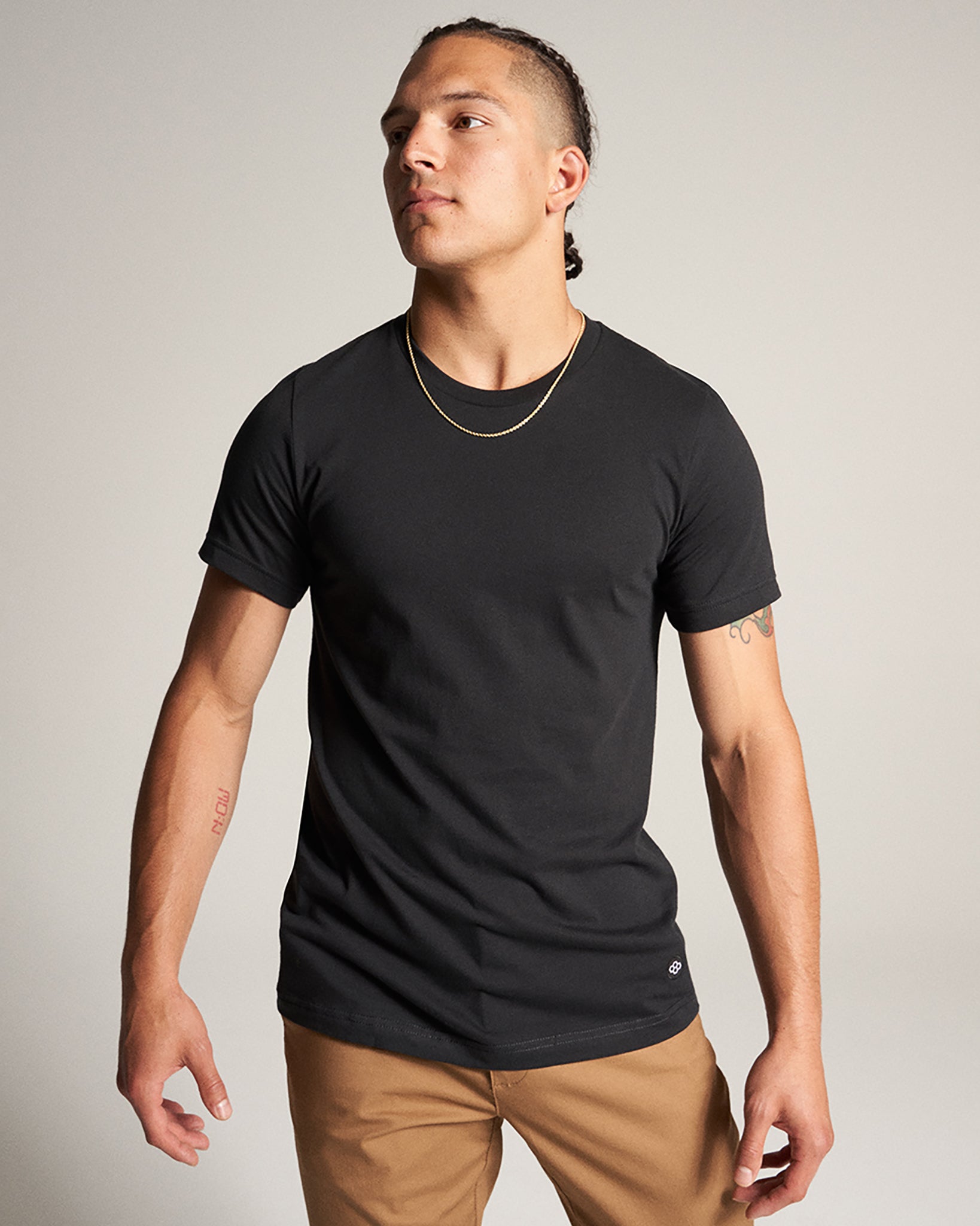 A young man poses confidently in a fitted black t-shirt and tan trousers complemented by a simple gold chain around his neck