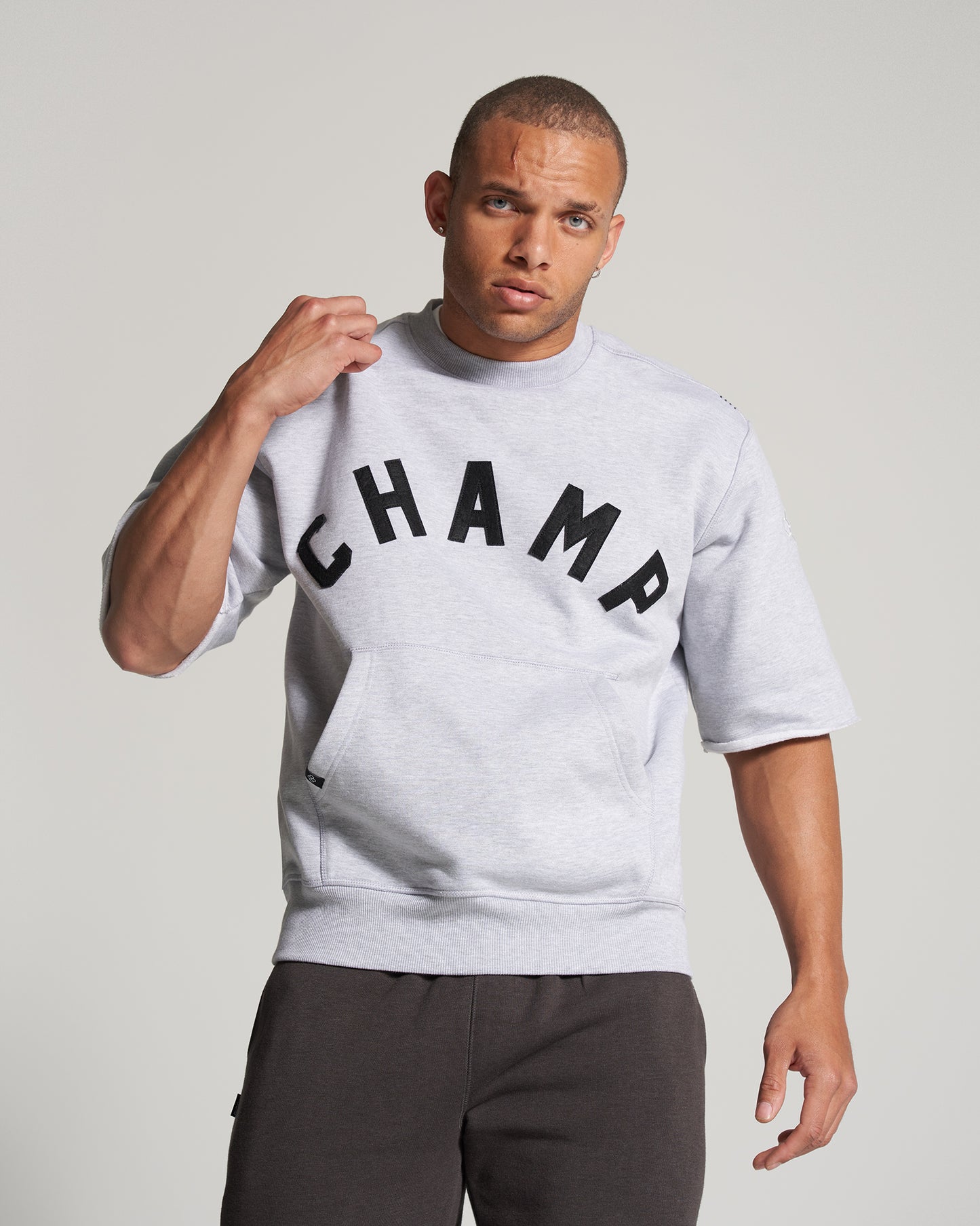 A young man wearing a light gray sweatshirt with CHAMP printed in bold black letters styled with casual black shorts posing confidently against a neutral background