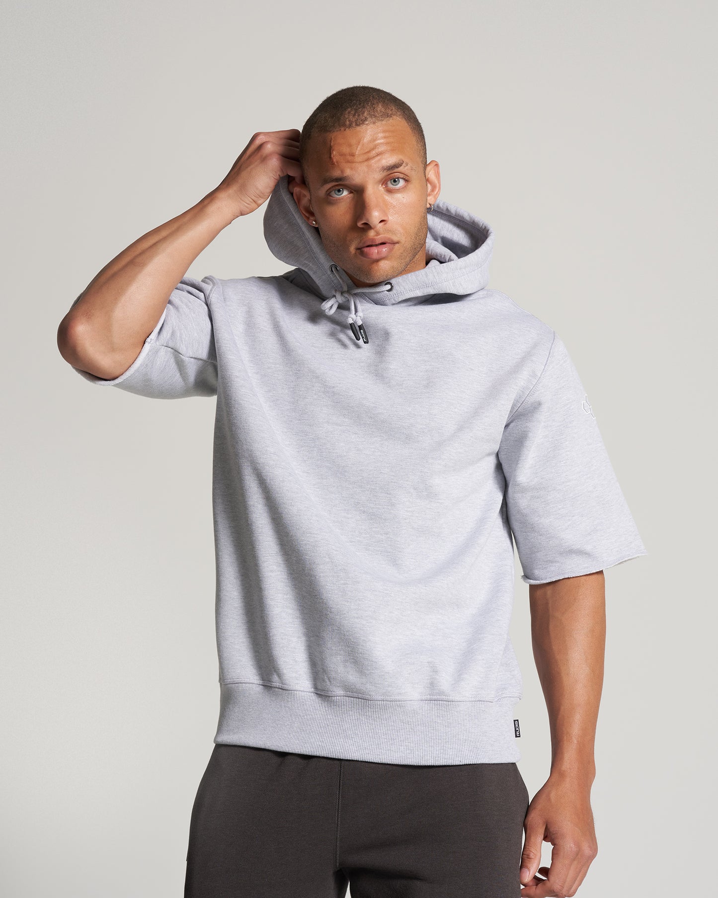 A man in a light gray hoodie is posing wearing a casual outfit that includes dark pants with a minimalist background enhancing his look