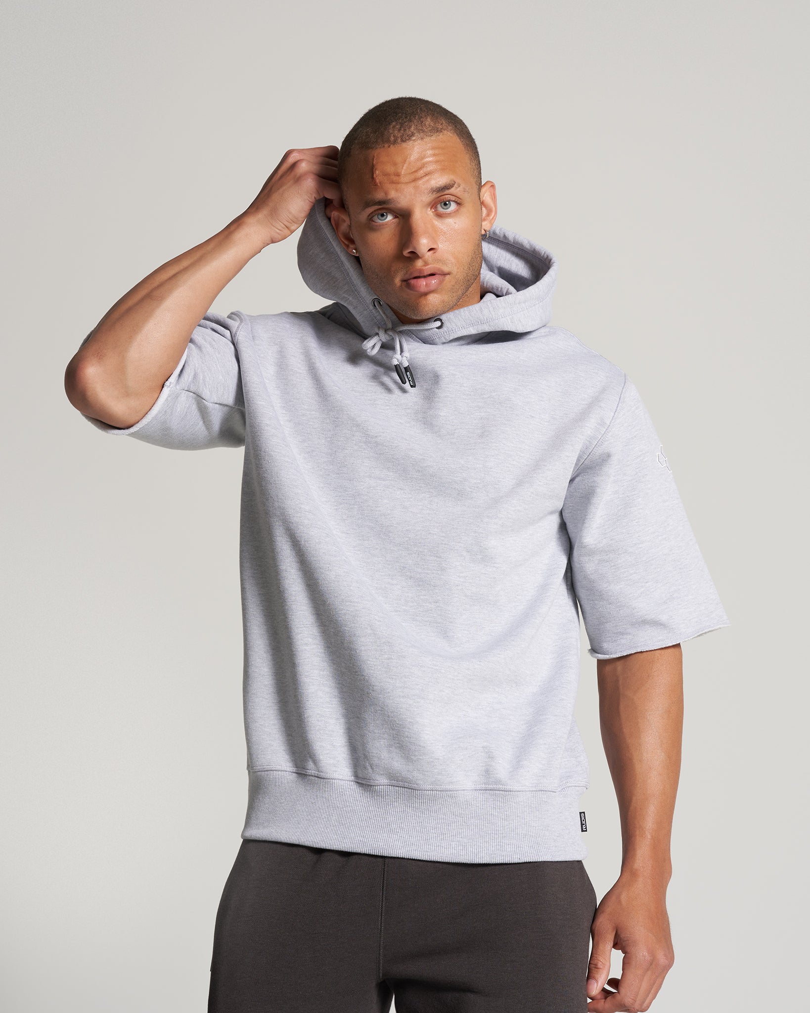 A man in a light gray hoodie is posing wearing a casual outfit that includes dark pants with a minimalist background enhancing his look