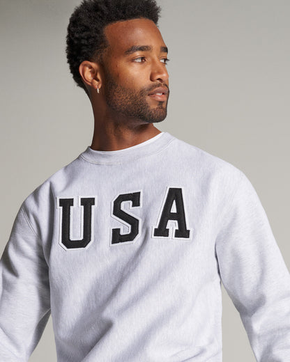 A man wearing a light gray sweatshirt featuring bold black letters spelling USA prominently across the chest showcasing a casual sporty look