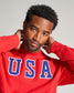 A confident young man in a red sweatshirt with USA emblazoned across the chest poses thoughtfully highlighting his modern style and casual demeanor