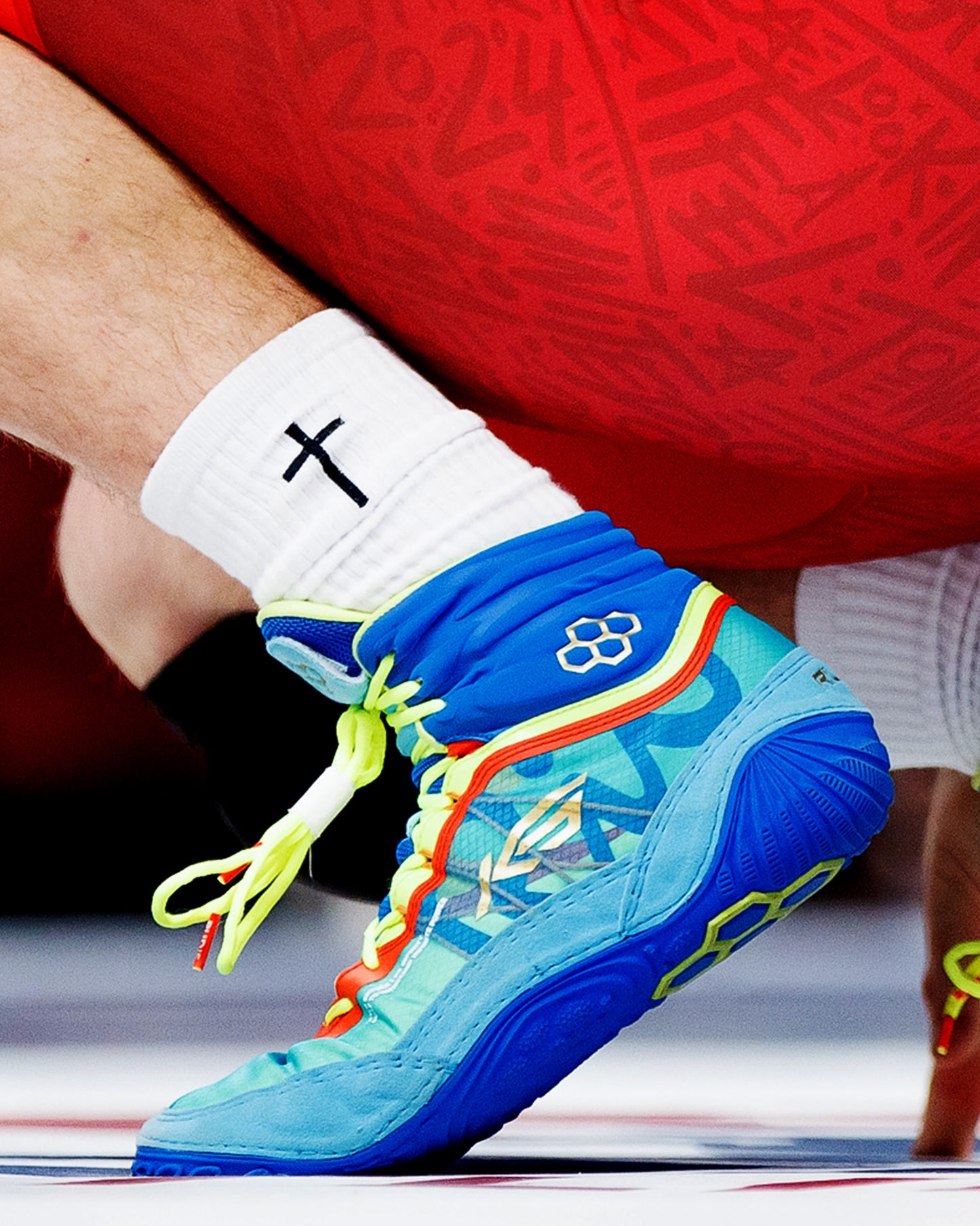 A vibrant blue and neon green wrestling shoe is being tied showcasing its unique design and color scheme against a background of red shorts and a white sock featuring a black cross