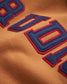 This close-up image features a textured fabric with bold stitched blue and red letters showcasing modern lettering on a warm orange-brown background