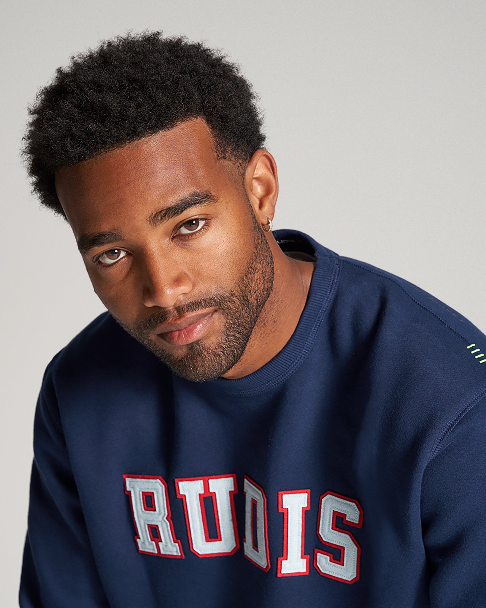 A man with short curly hair is wearing a navy sweatshirt featuring the word RUDIS in bold colorful lettering with a neutral gray background