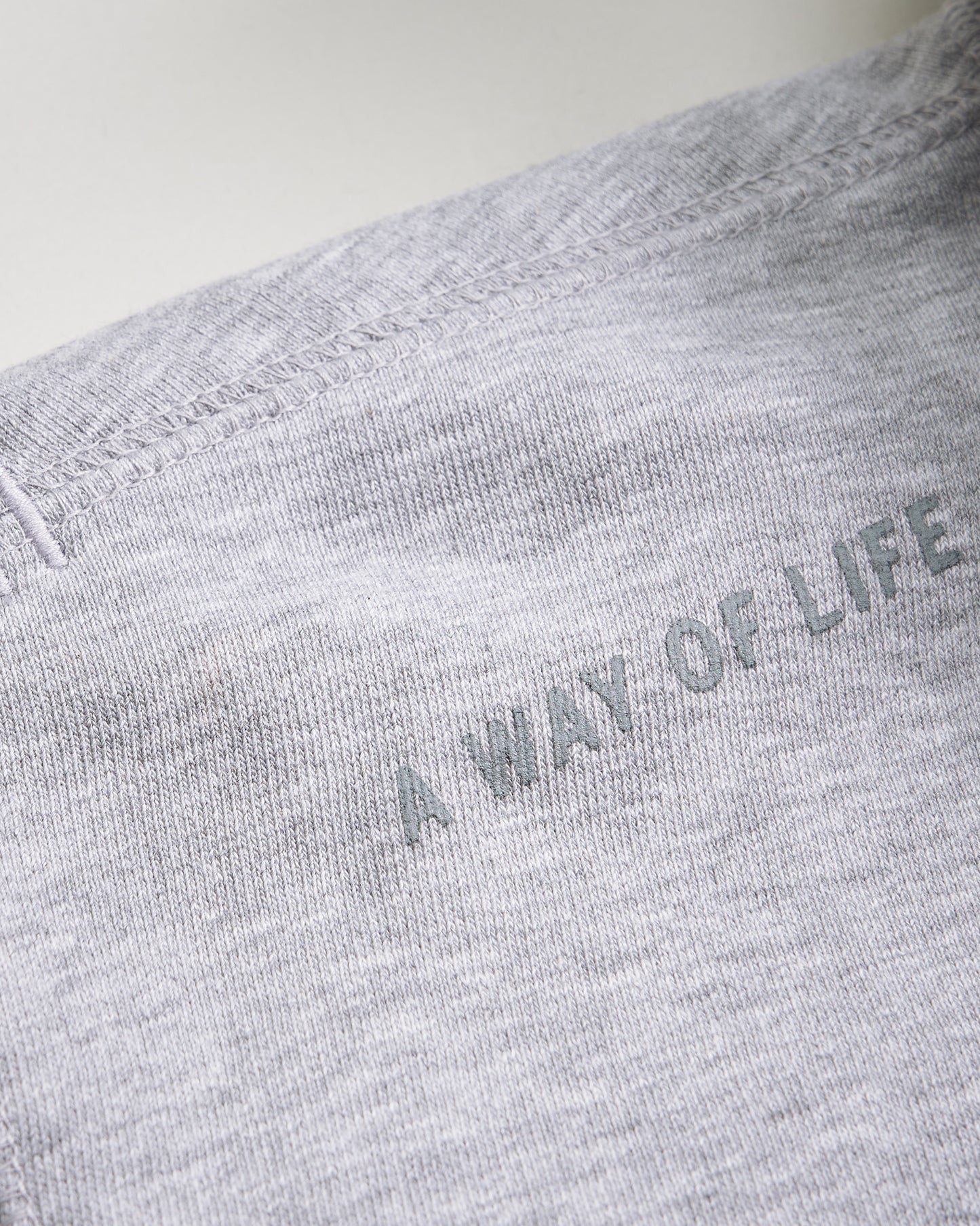 A close-up view of a light gray fabric with the phrase A WAY OF LIFE printed in subtle green text highlighting the texture and stitching details of the garment