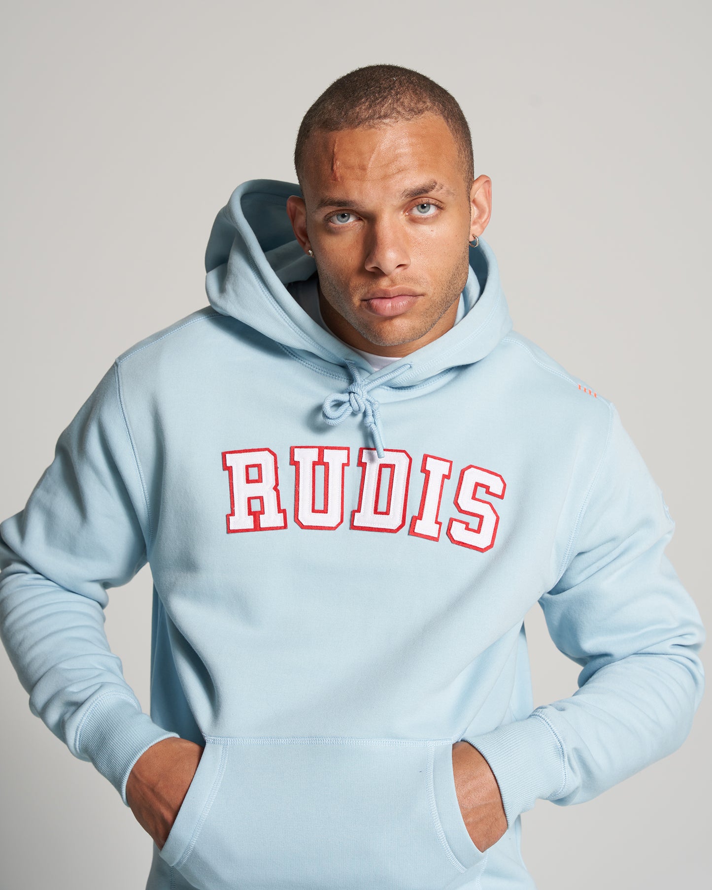 A male model in a light blue hoodie with RUDIS printed in bold red letters striking a serious expression with his hands in his pockets