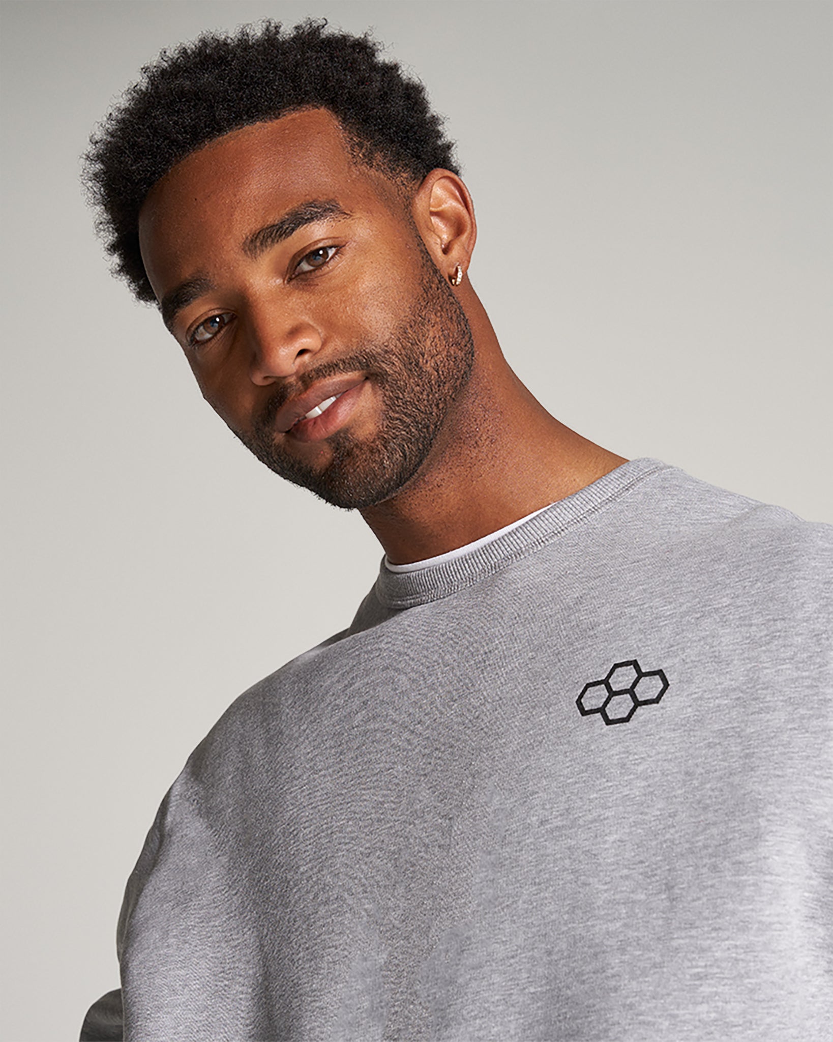 A close-up portrait of a smiling man wearing a gray sweatshirt with an embroidered logo on the chest showcasing his stylish hairstyle and confident expression