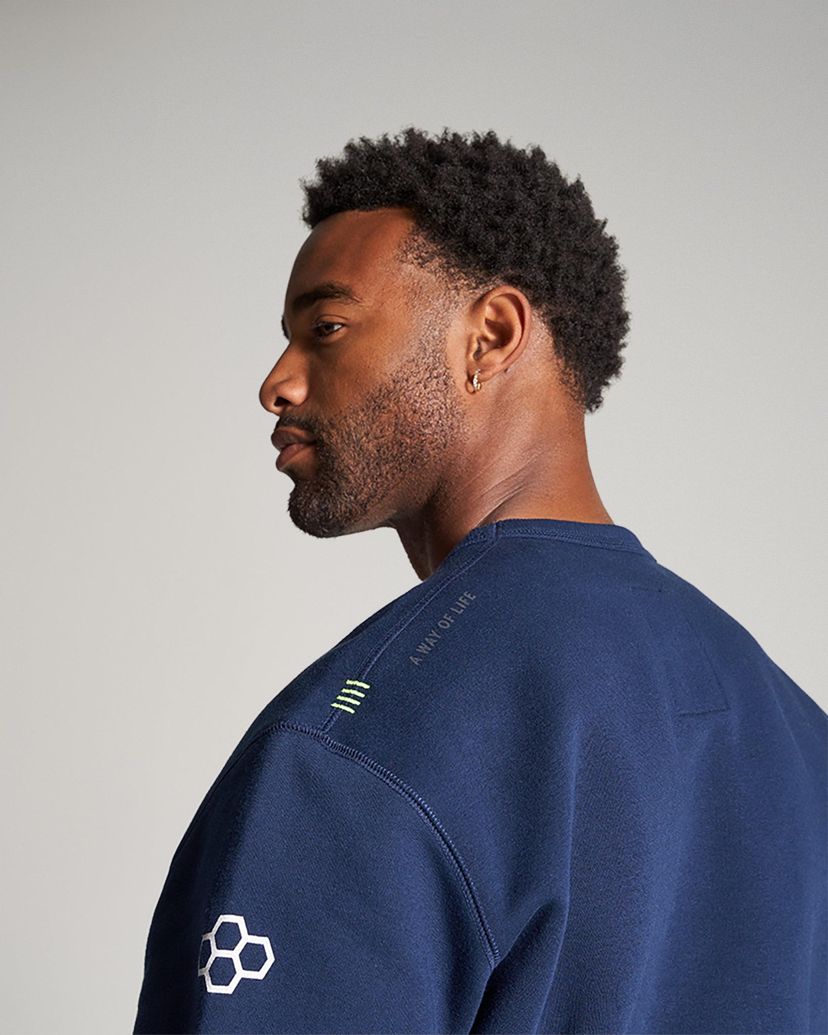 A man with a well-groomed beard wears a navy blue sweatshirt featuring unique detailing on the shoulders and an emblem on the sleeve