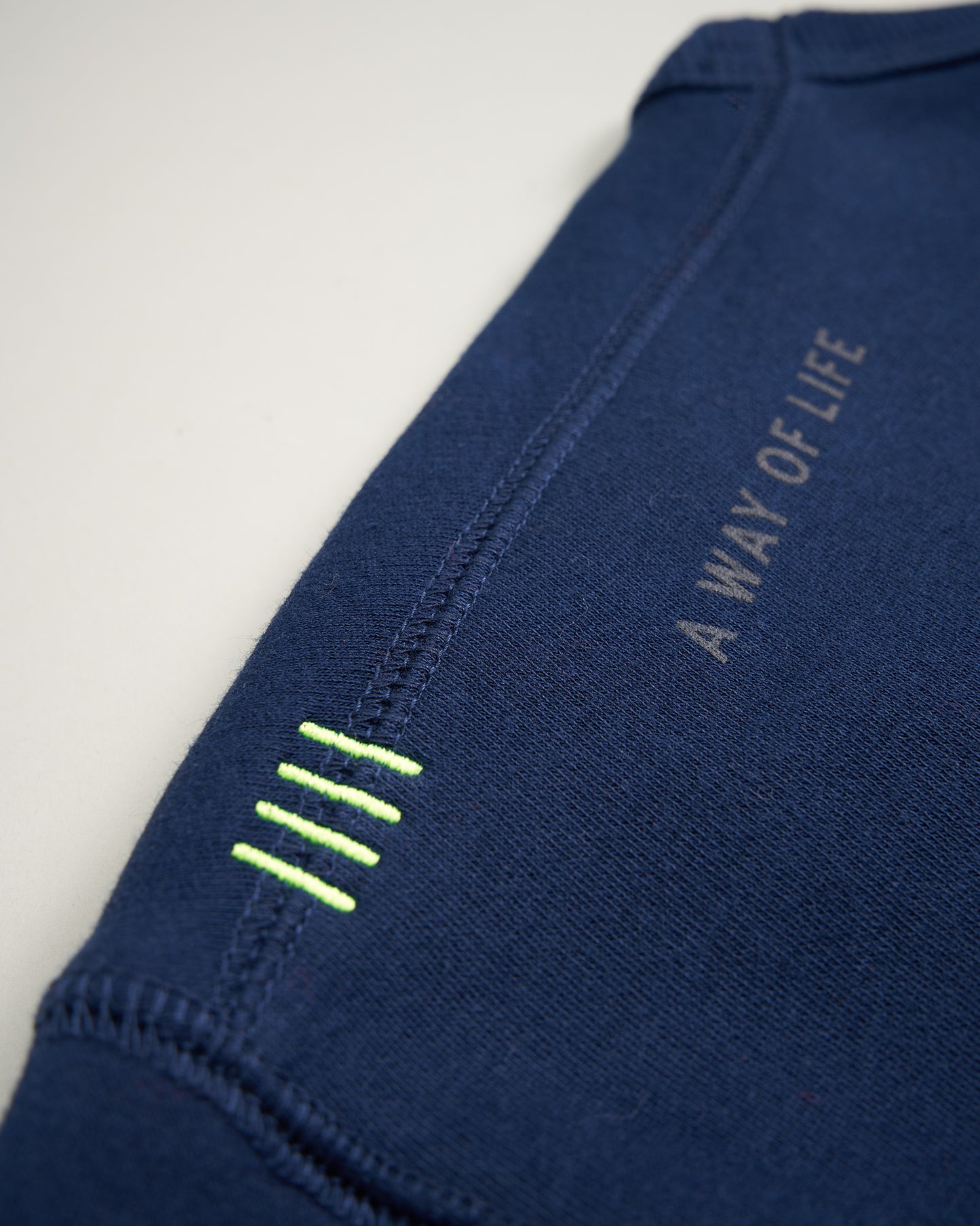 A close-up of a navy blue garment featuring a stitched label that reads A WAY OF LIFE and decorative neon yellow stitching on the sleeve