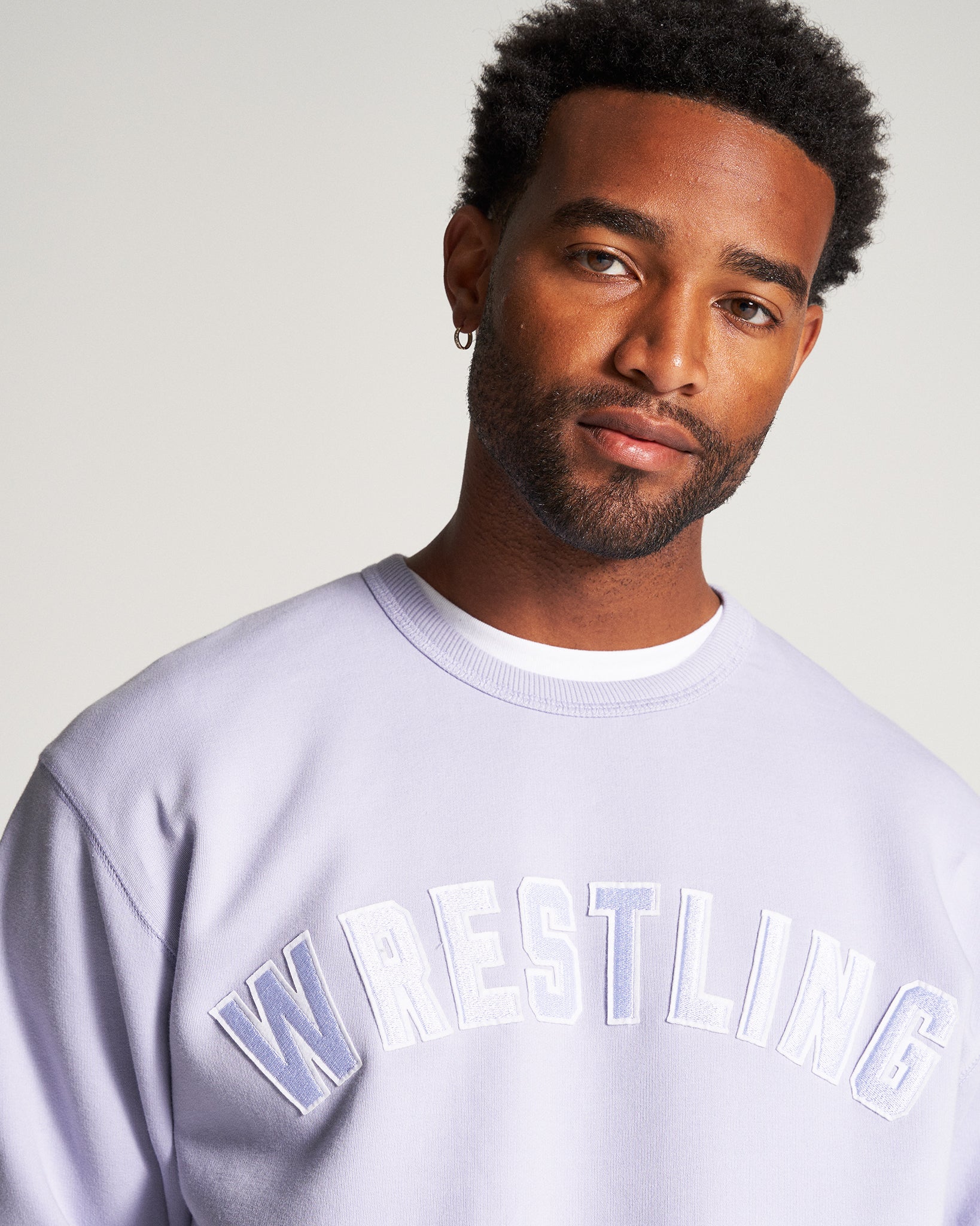 A man with a stylish short hairstyle wearing a lavender sweatshirt featuring WRESTLING in bold text