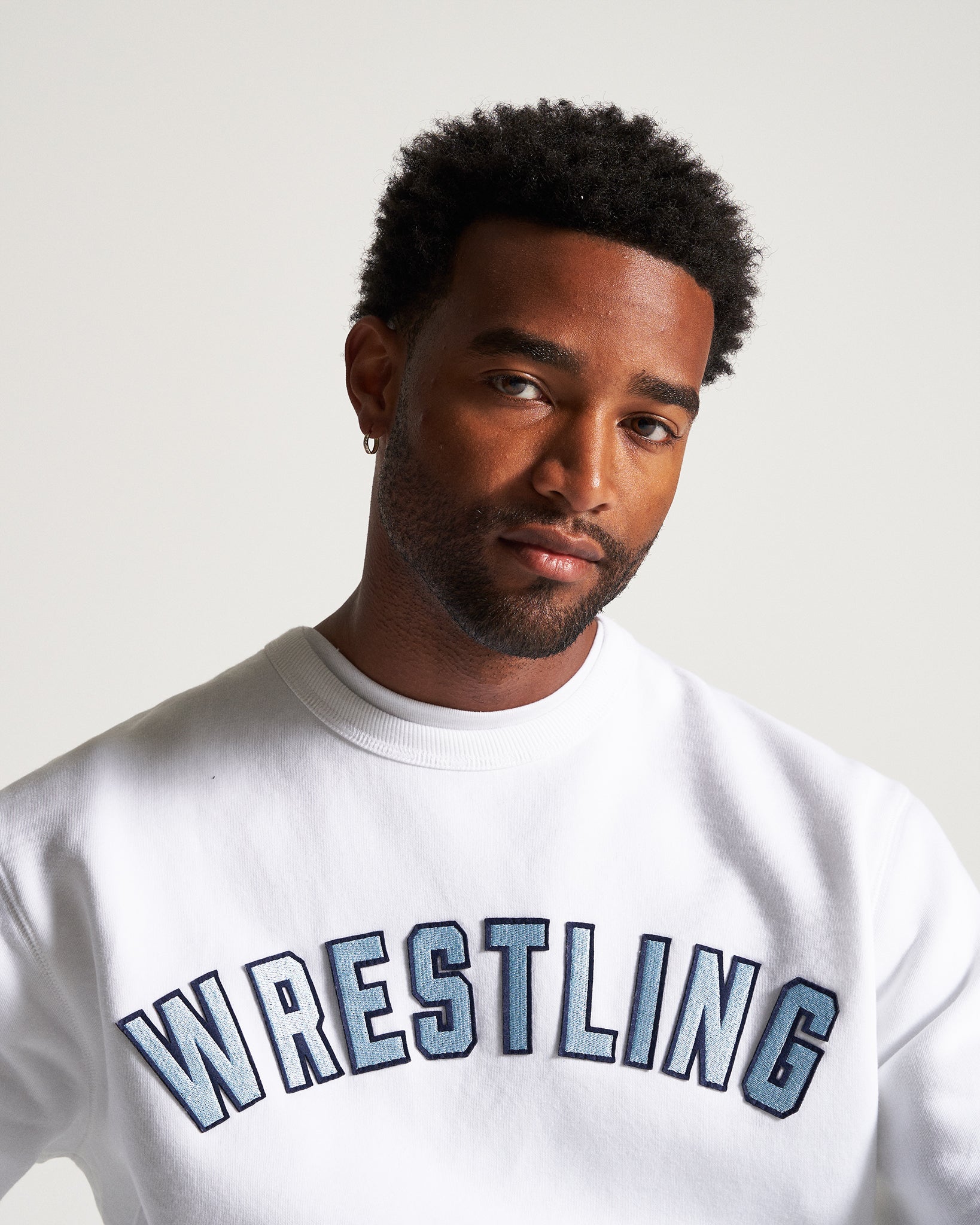 A man with a close-cropped beard and textured hair is wearing a white sweatshirt with the word WRESTLING prominently displayed in bold blue embroidery