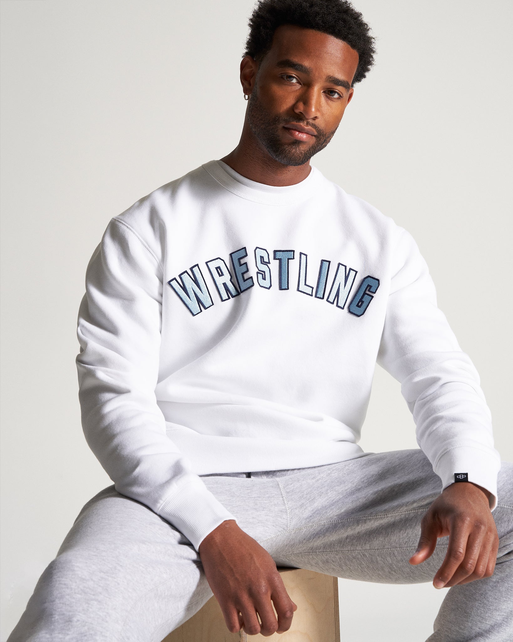 A man poses in a white sweatshirt with WRESTLING printed prominently on the front sitting casually on a wooden block while wearing gray sweatpants