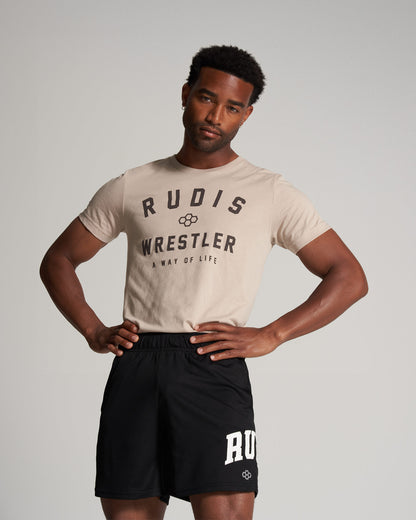 A young man poses confidently in a beige t-shirt and black athletic shorts featuring the brand RUDIS and the word WRESTLER