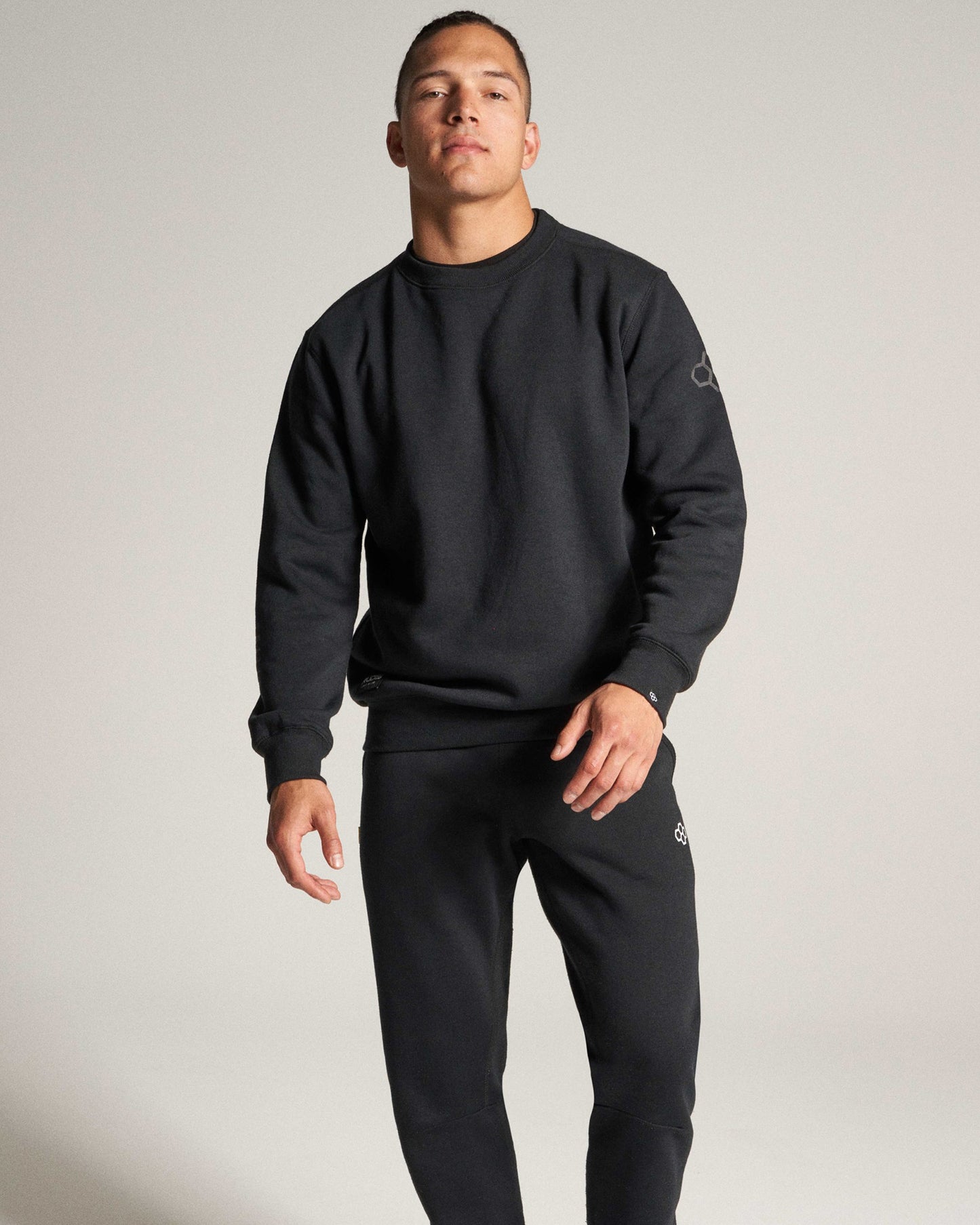 A man stands confidently in a fitted black sweatshirt and matching sweatpants set against a minimalist gray background