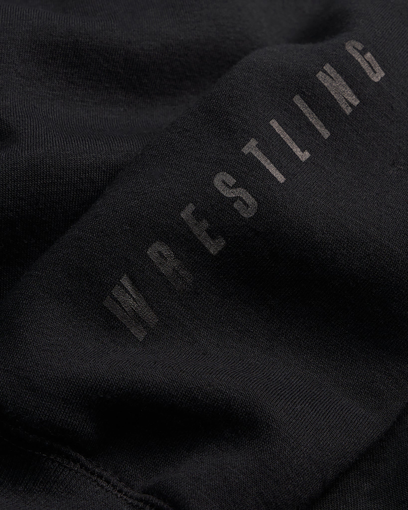 This close-up image features a black fabric with the word WRESTLING printed in a tonal style emphasizing its texture and minimalist design