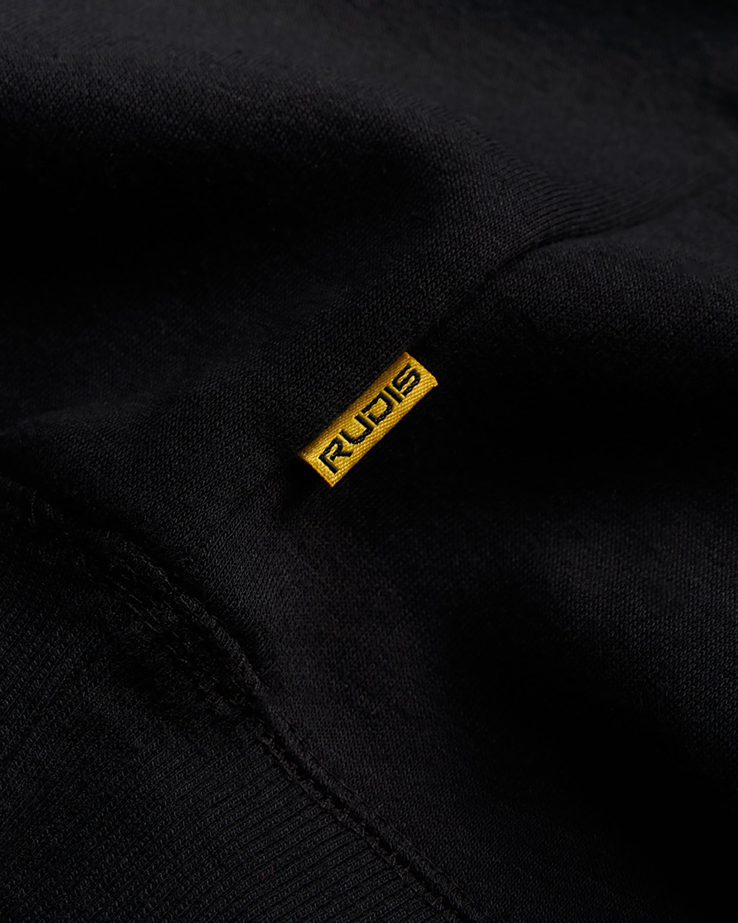 A close-up view of a black fabric with a distinct yellow tag featuring the word RUDIS