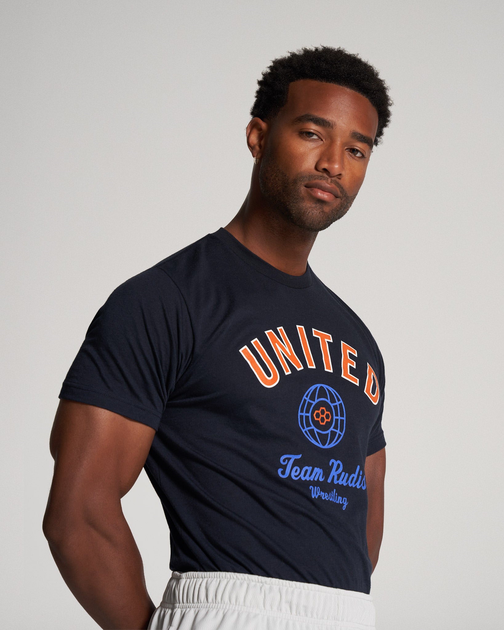 A confident young man poses in a navy blue t-shirt featuring bold graphics and lettering promoting a wrestling team