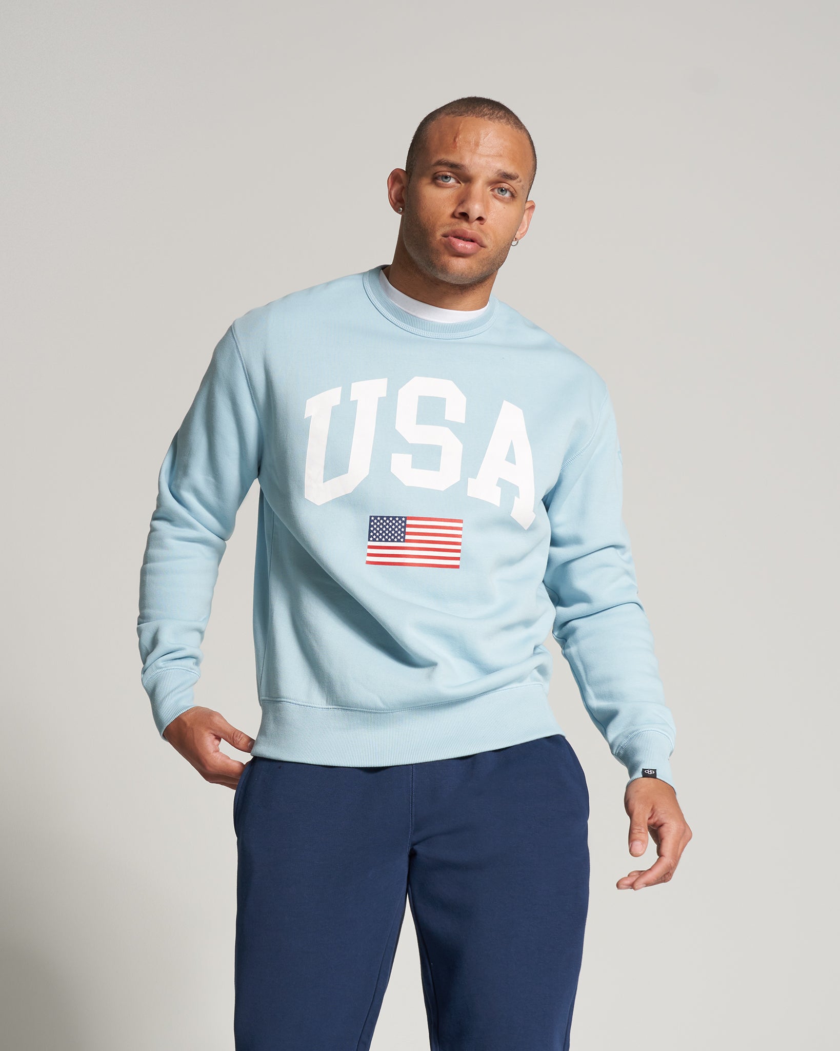 A man wearing a light blue sweatshirt with USA printed on it and an American flag emblem paired with dark pants stands against a neutral backdrop