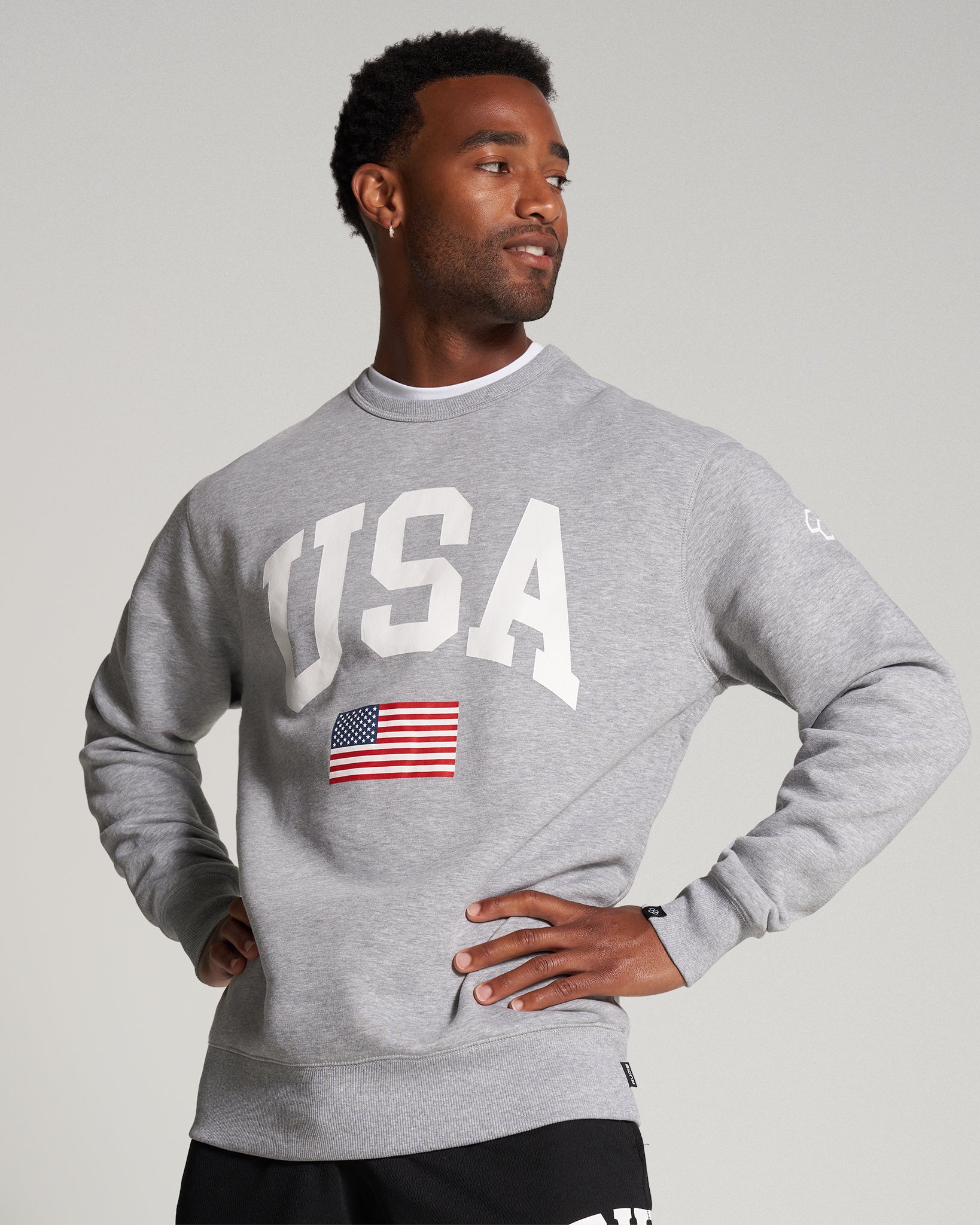 A man wearing a light gray sweatshirt featuring USA in bold letters and an American flag posing confidently with a casual expression