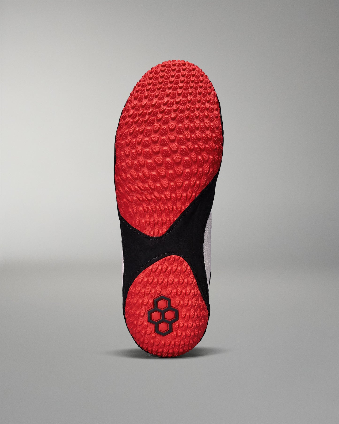 The underside of a shoe showcases a vibrant red textured outsole with a traction pattern and a black section featuring a hexagonal logo, set against a muted gray background.