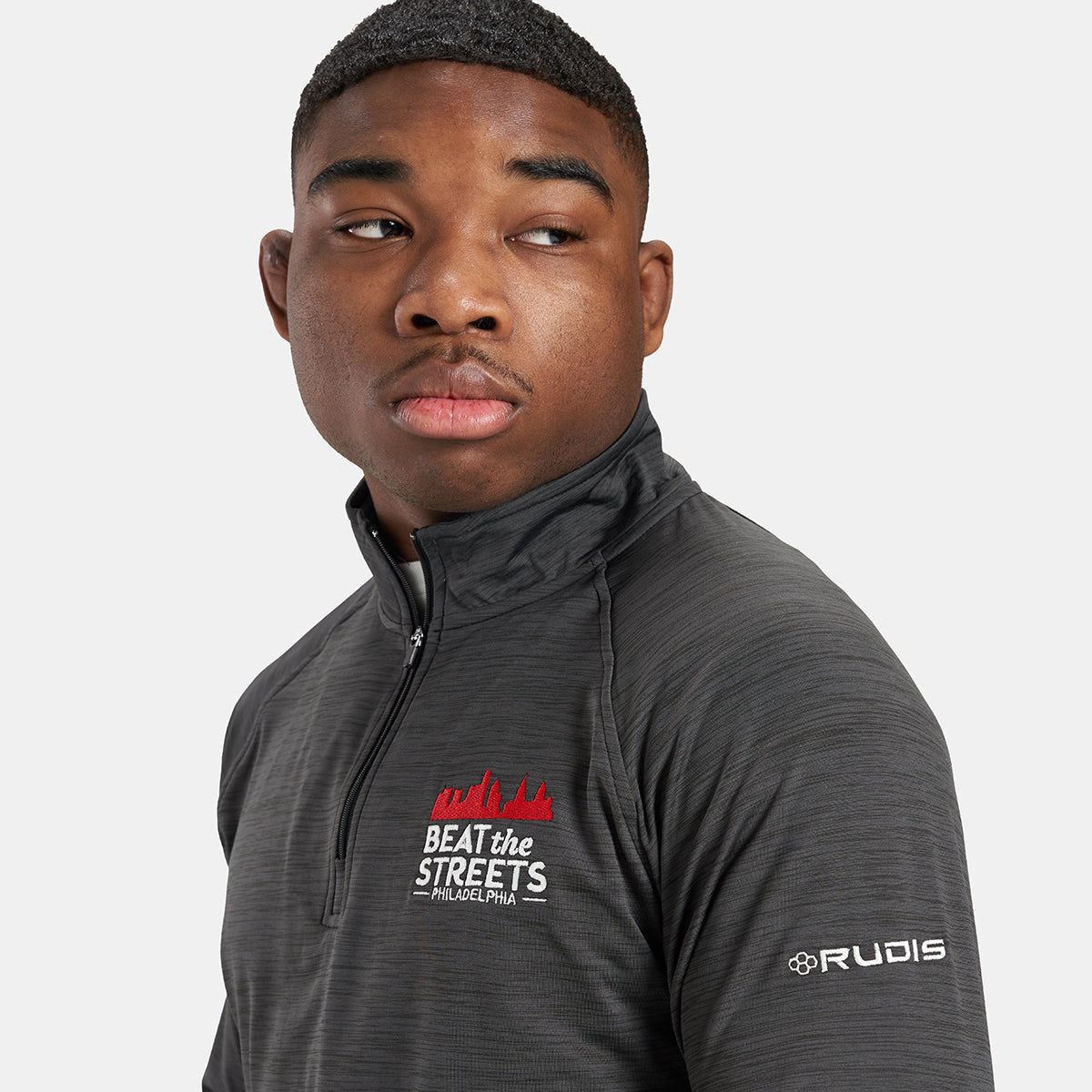 A young man wearing a black athletic half-zip shirt with BEAT the STREETS PHILADELPHIA logo and RUDIS branding on the sleeve facing sideways with a confident expression