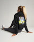 A woman is sitting on the floor in a stylish cropped black hoodie featuring a colorful graphic design paired with gray leggings and white athletic shoes