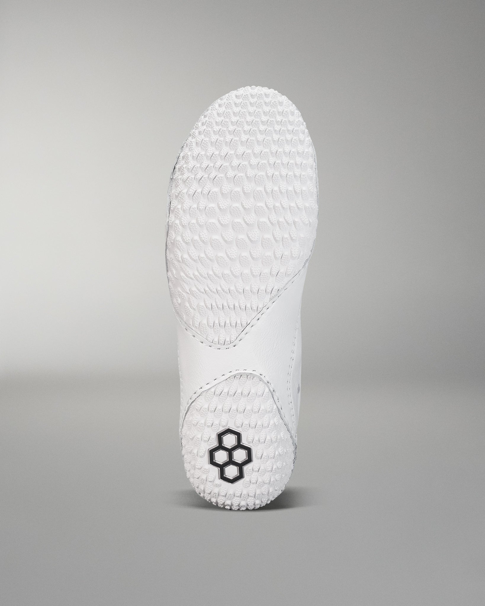 The sole of a white shoe, featuring a textured grip surface and a small black hexagonal logo, is set against a soft gray background.