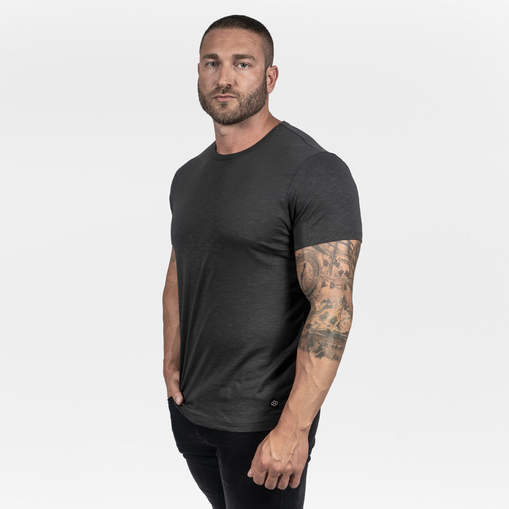 A muscular man stands confidently wearing a fitted dark gray t-shirt and black jeans showcasing his tattoos and strong physique against a plain white background