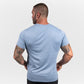 A muscular man stands with his back to the camera wearing a light blue t-shirt and black pants showcasing his tattoos and athletic build against a minimalist gray background