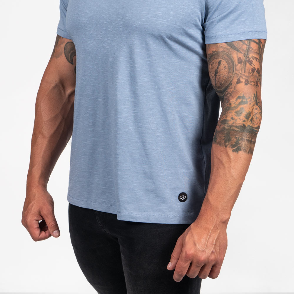 A muscular man is wearing a light blue t-shirt paired with dark jeans showcasing his well-defined arms and tattoos emphasizing a fitness-oriented style