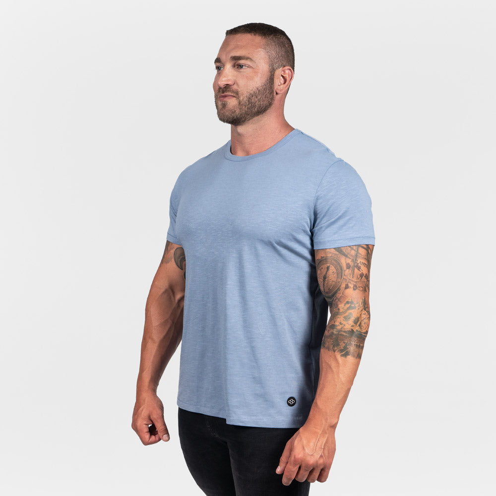 This image features a muscular man in a light blue t-shirt and black jeans showcasing a confident pose against a plain white background