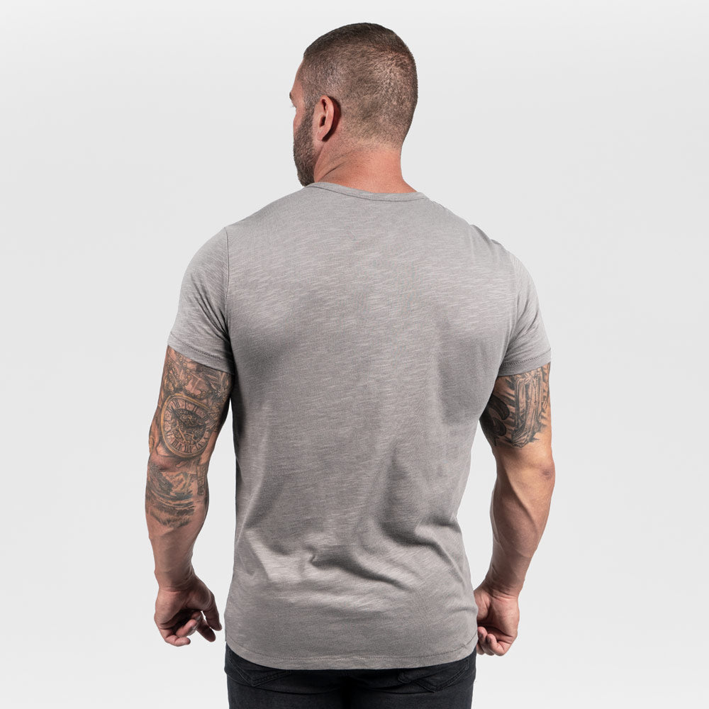 This image features a muscular man viewed from the back wearing a fitted gray t-shirt showcasing detailed tattoos on his arms against a neutral background