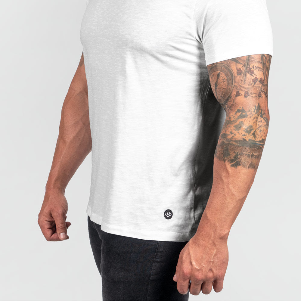 A close-up of a muscular mans arm wearing a fitted white t-shirt showcasing intricate tattoos and a branded tag near the hem