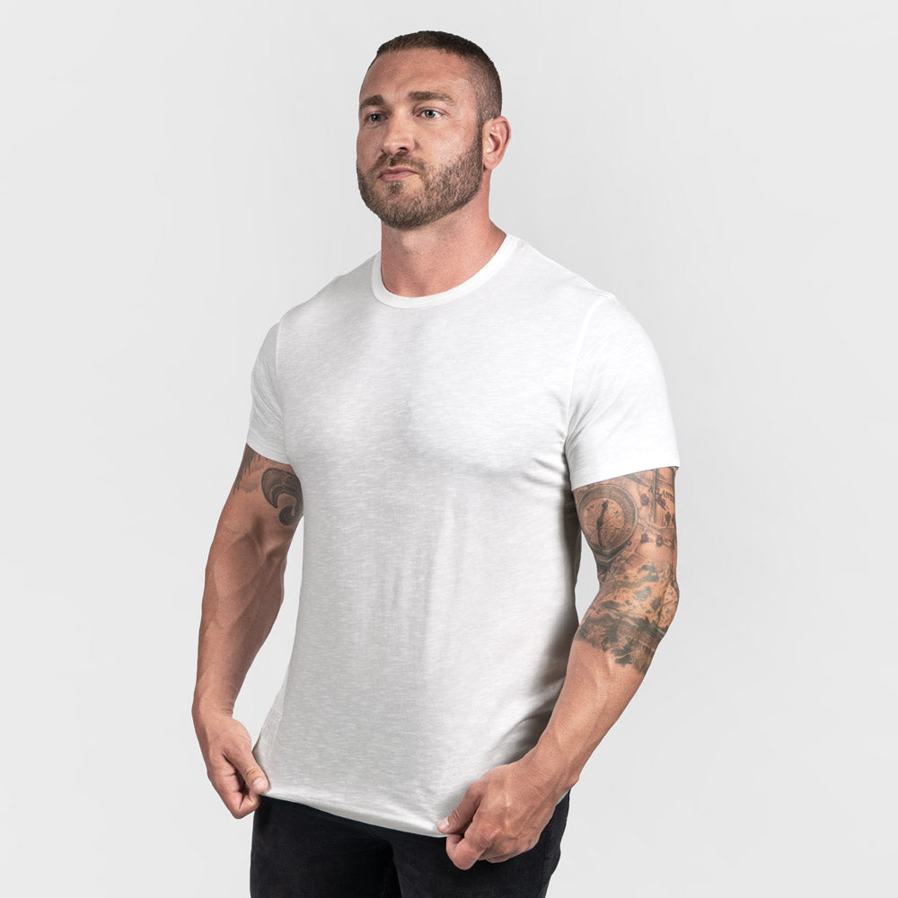 A muscular man with a beard stands confidently wearing a fitted white t-shirt against a light gray background