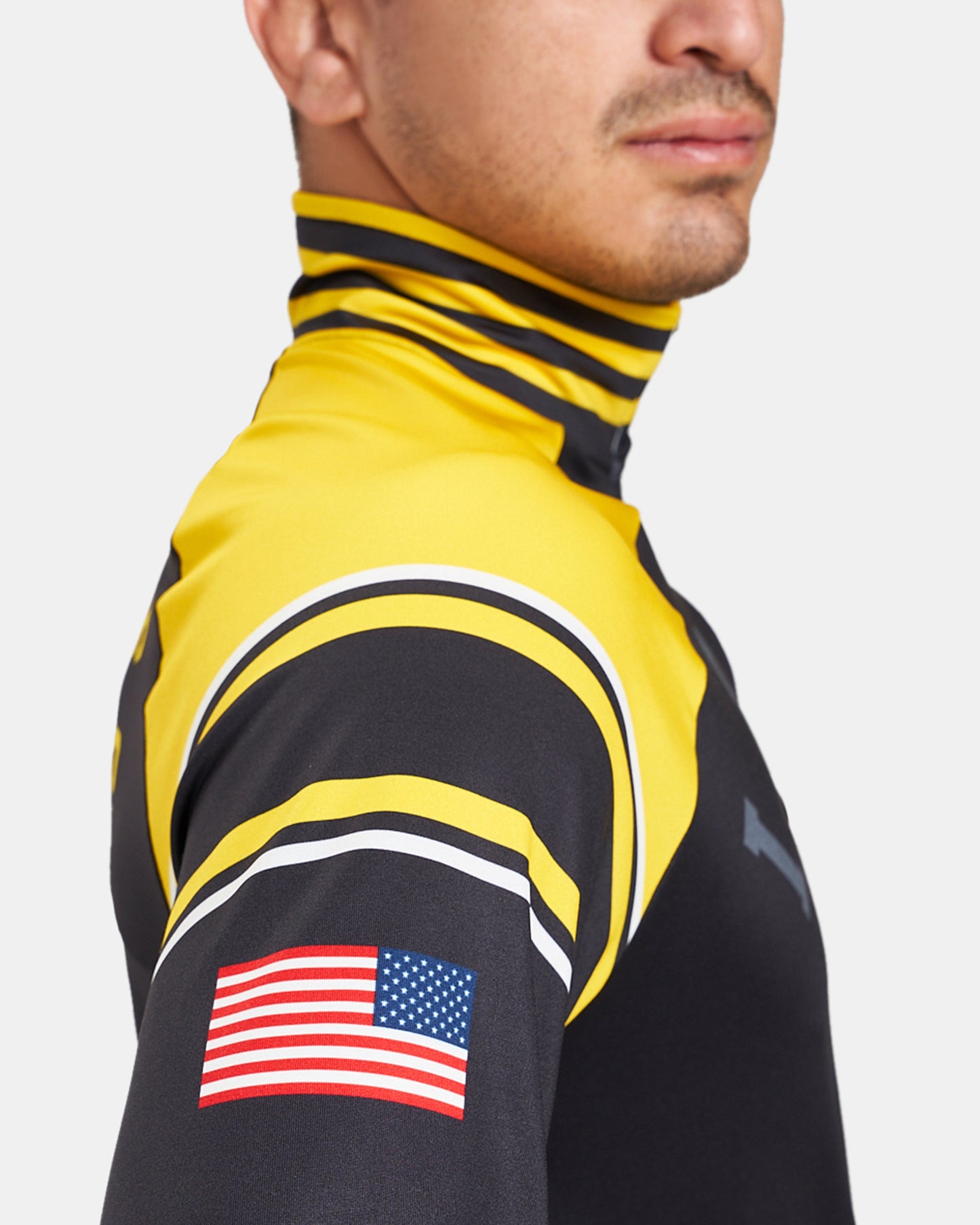 A man in a vibrant yellow high-collared athletic shirt with black and white stripes featuring an American flag patch on the sleeve confidently poses to showcase the apparels design and patriotic elements