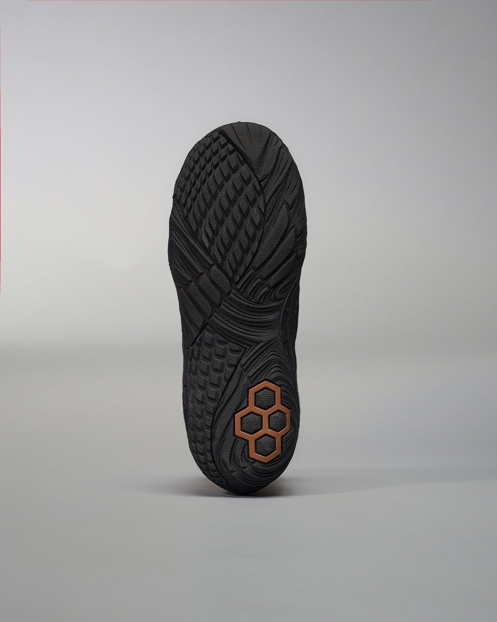 The image displays the textured sole of a black athletic shoe highlighting its unique tread pattern and distinctive hexagonal logo in bronze at the heel