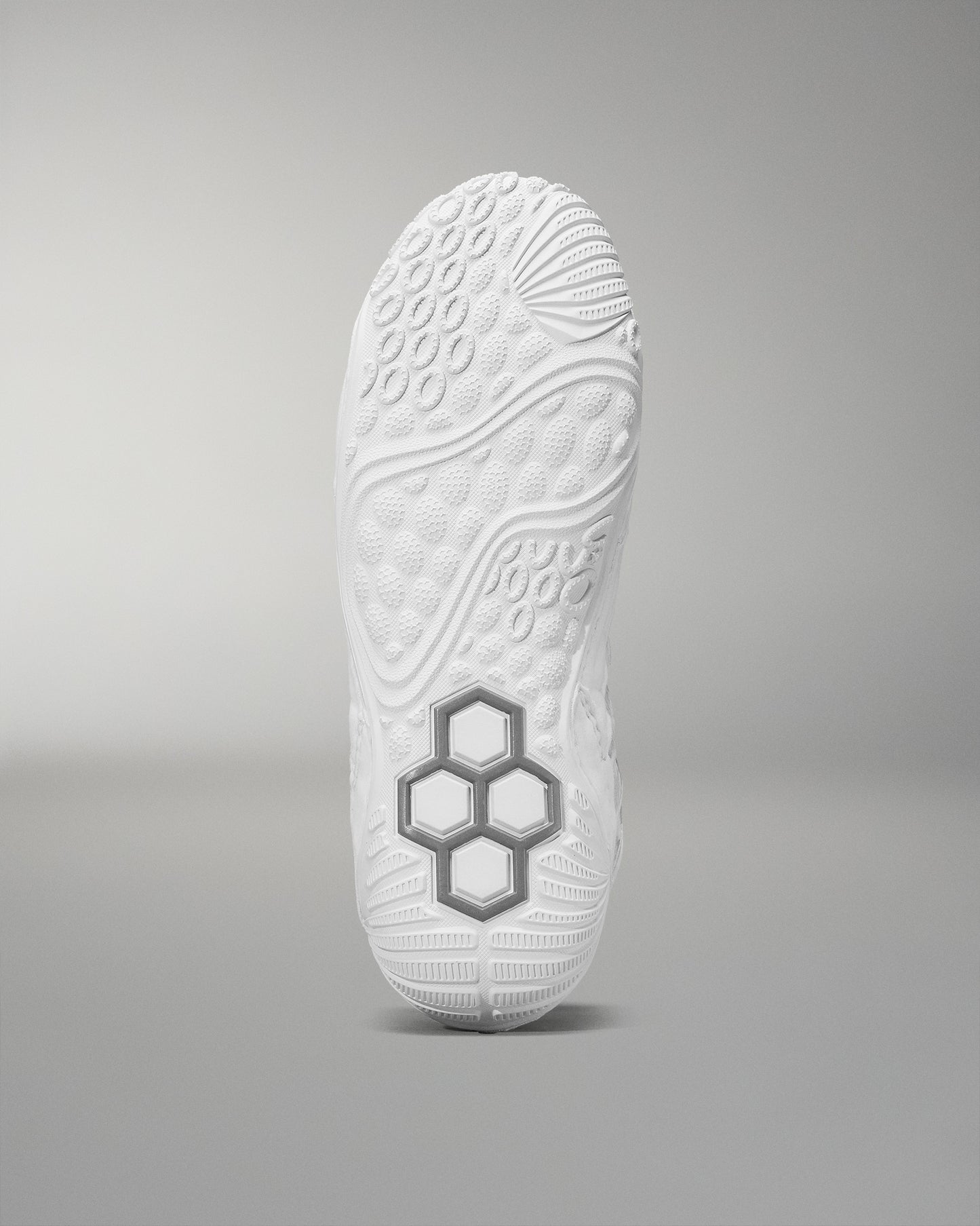 The sole of a white athletic shoe, set against a soft gray background, showcases a textured design with raised patterns and a prominent gray hexagonal structure for improved grip.