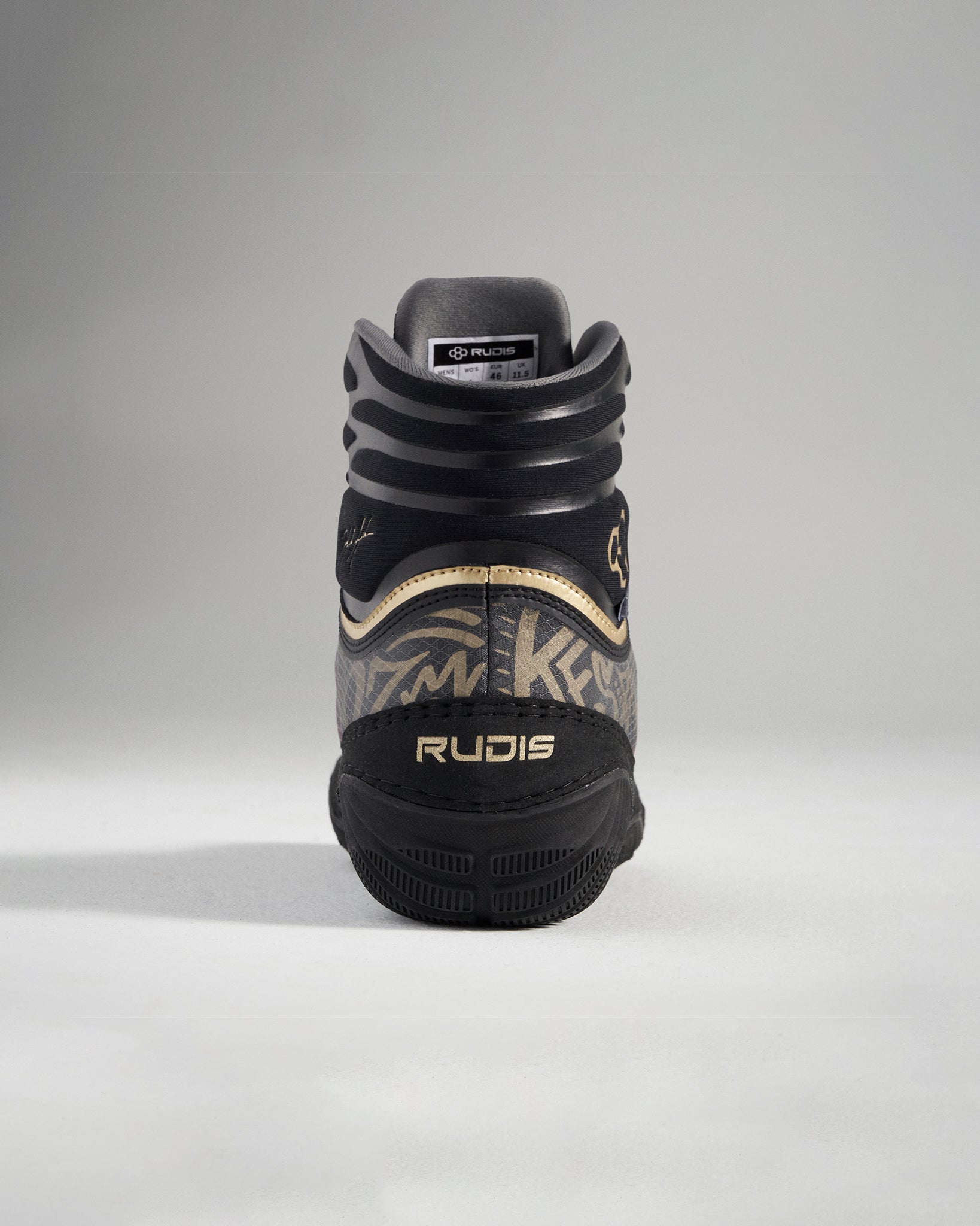 A rear view of a black and gold performance shoe designed for wrestling featuring a textured pattern and the brand name RUDIS prominently displayed