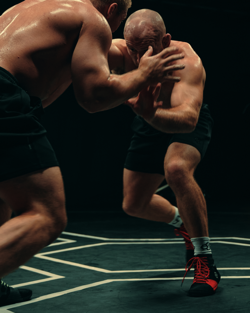 Two shirtless wrestlers engage intensely on a dark mat with white lines, highlighting athleticism and focus.