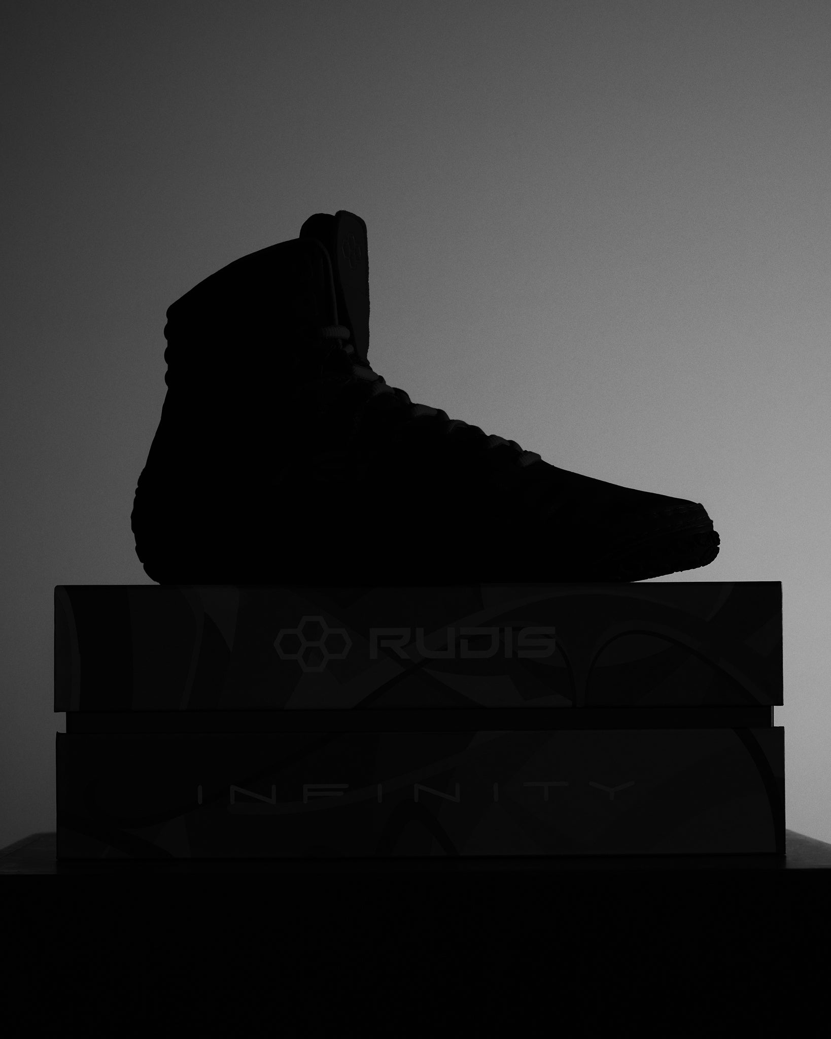 A black athletic shoe silhouette atop boxes labeled 'RUDIS' and 'INFINITY', showcased in a minimalist and illuminated setting.