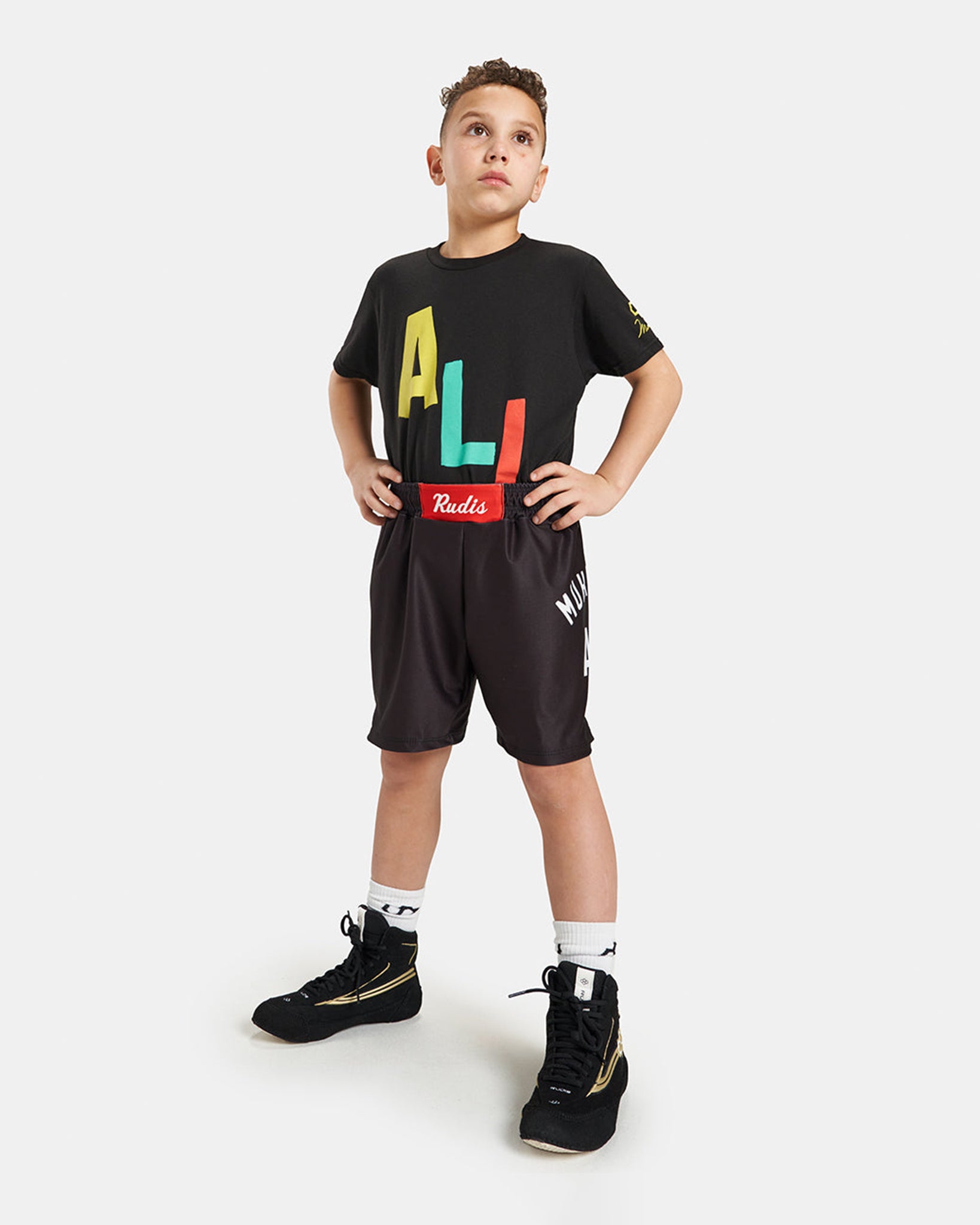 A young boy wearing a colorful graphic t-shirt and black athletic shorts poses confidently in stylish sports shoes