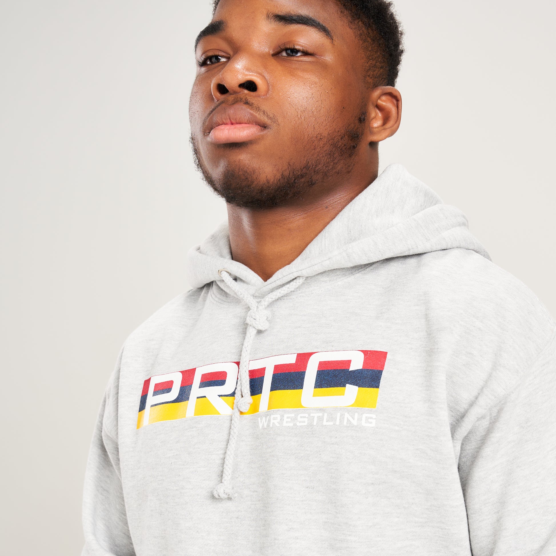 A young man poses confidently in a light gray hoodie featuring PRTC WRESTLING printed in bold colors on the front standing against a neutral background