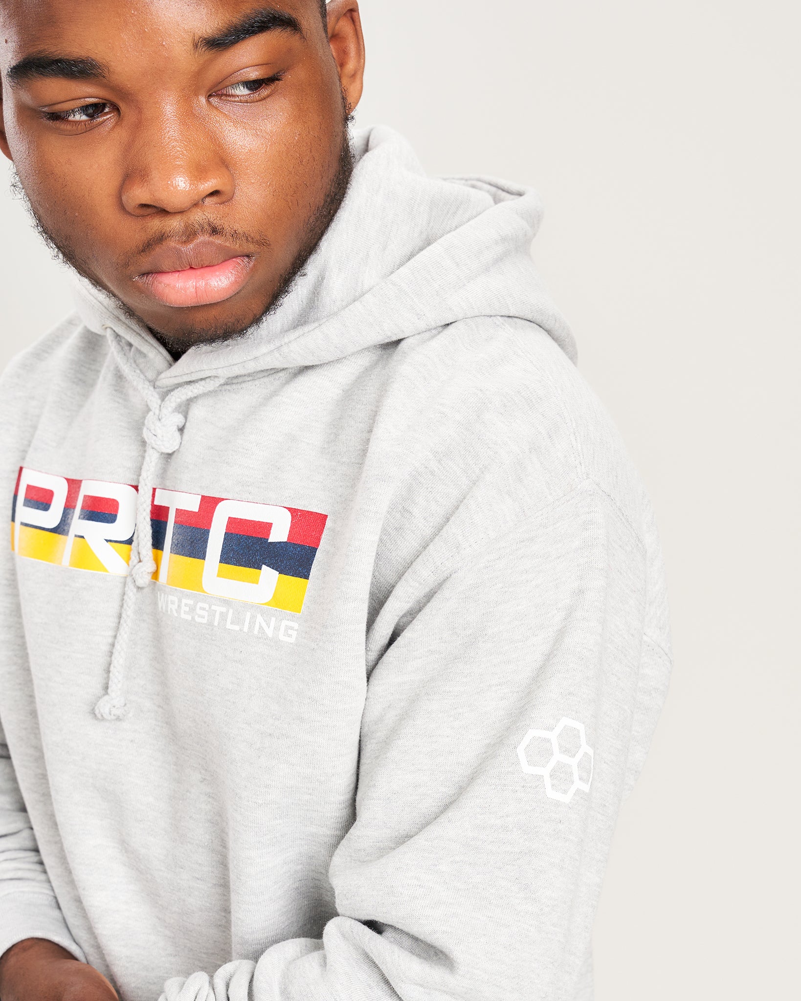 A young person wearing a light gray hoodie with a colorful PRTC Wrestling logo and a subtle design on the sleeve showcasing a thoughtful expression against a neutral background