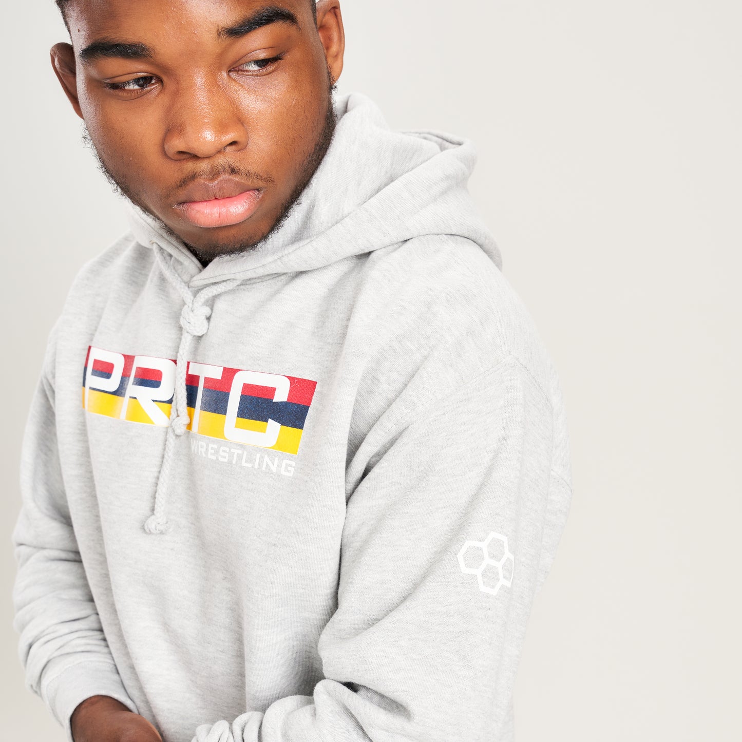 A young man wearing a light gray hoodie with a colorful PRTC Wrestling logo looking thoughtfully to the side against a neutral backdrop