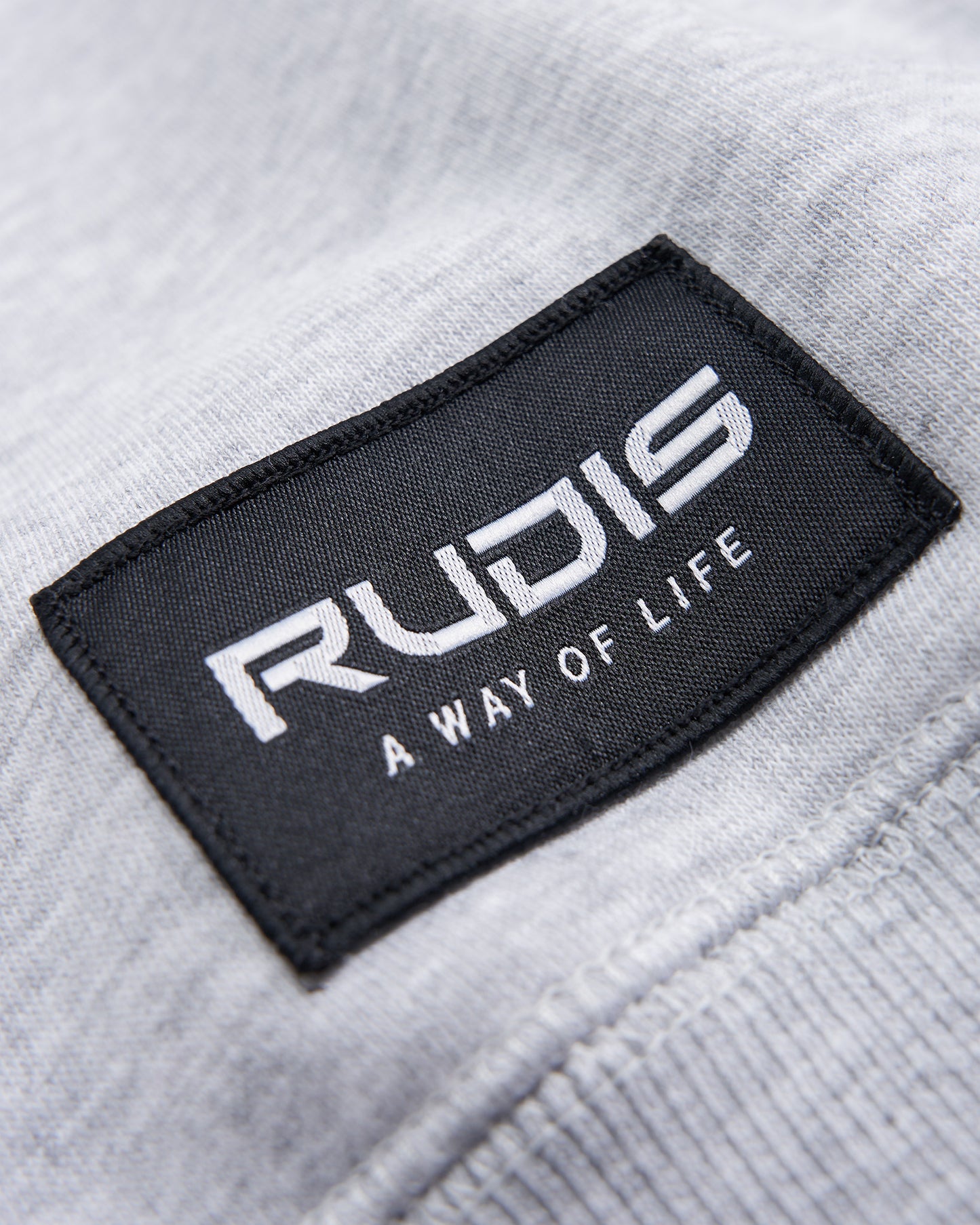 A close-up view of a black fabric label featuring the word RUDIS and the phrase A WAY OF LIFE on a light gray textured garment