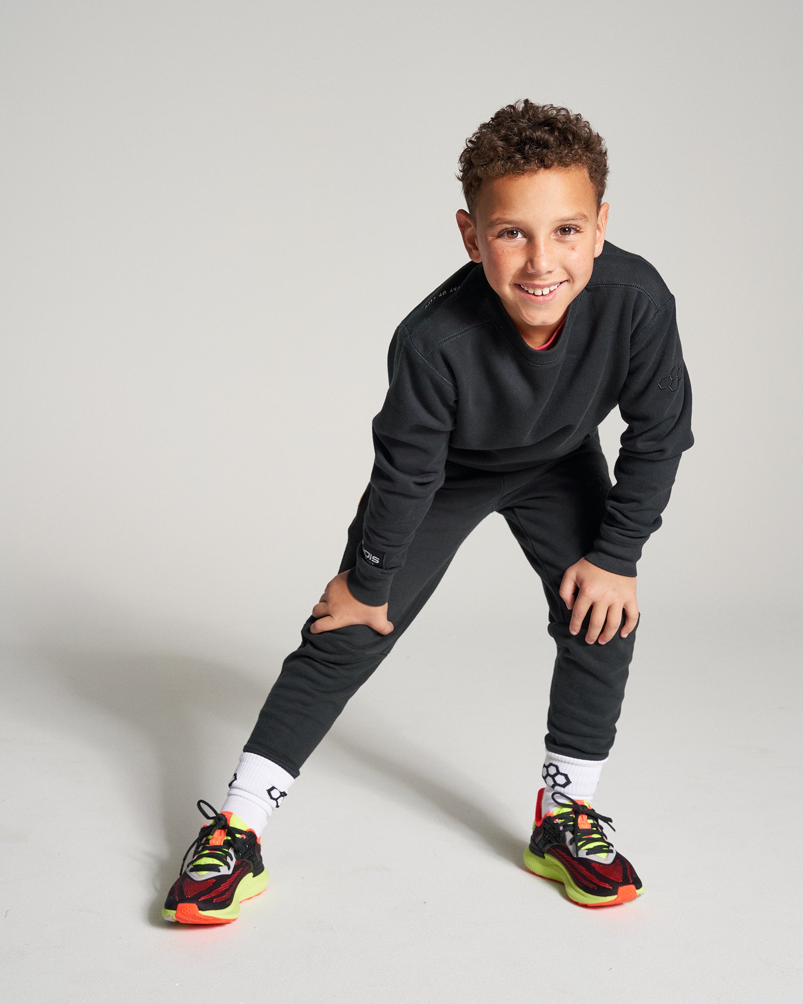 A young boy in stylish athletic wear poses playfully in a studio setting showcasing his vibrant sneakers and cheerful demeanor