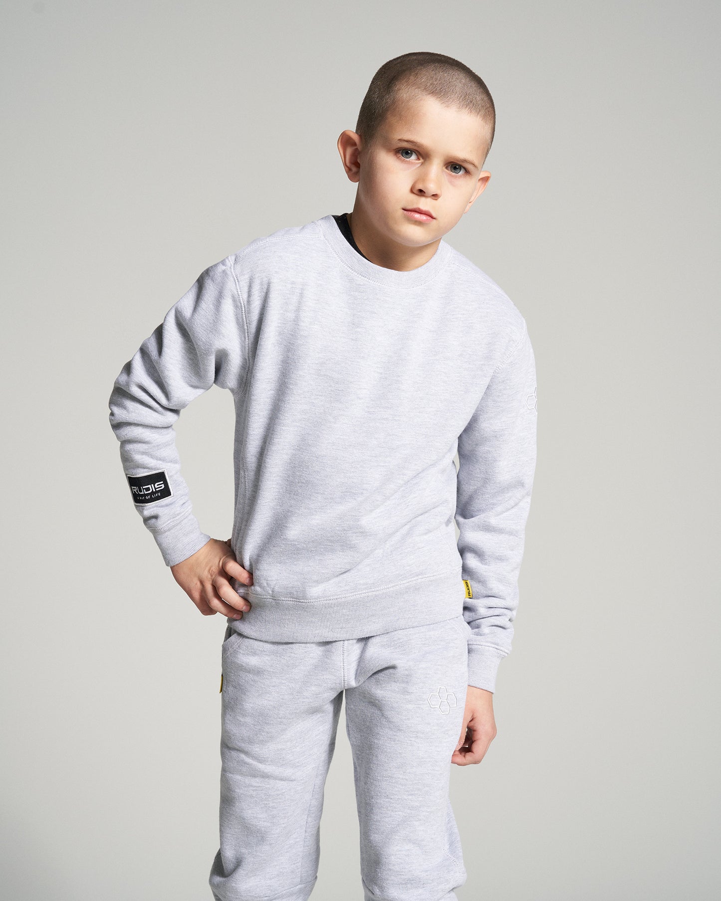 A young boy poses in a stylish gray tracksuit featuring a crew neck sweatshirt and matching sweatpants set against a simple gray background