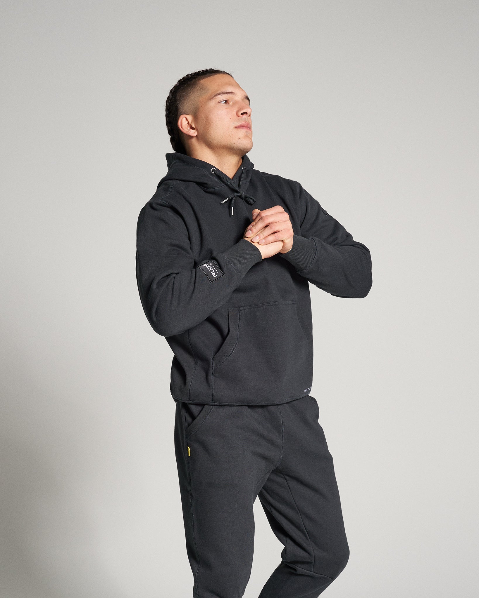 A man in a black hoodie and sweatpants strikes a thoughtful pose against a plain backdrop exuding a casual yet stylish vibe